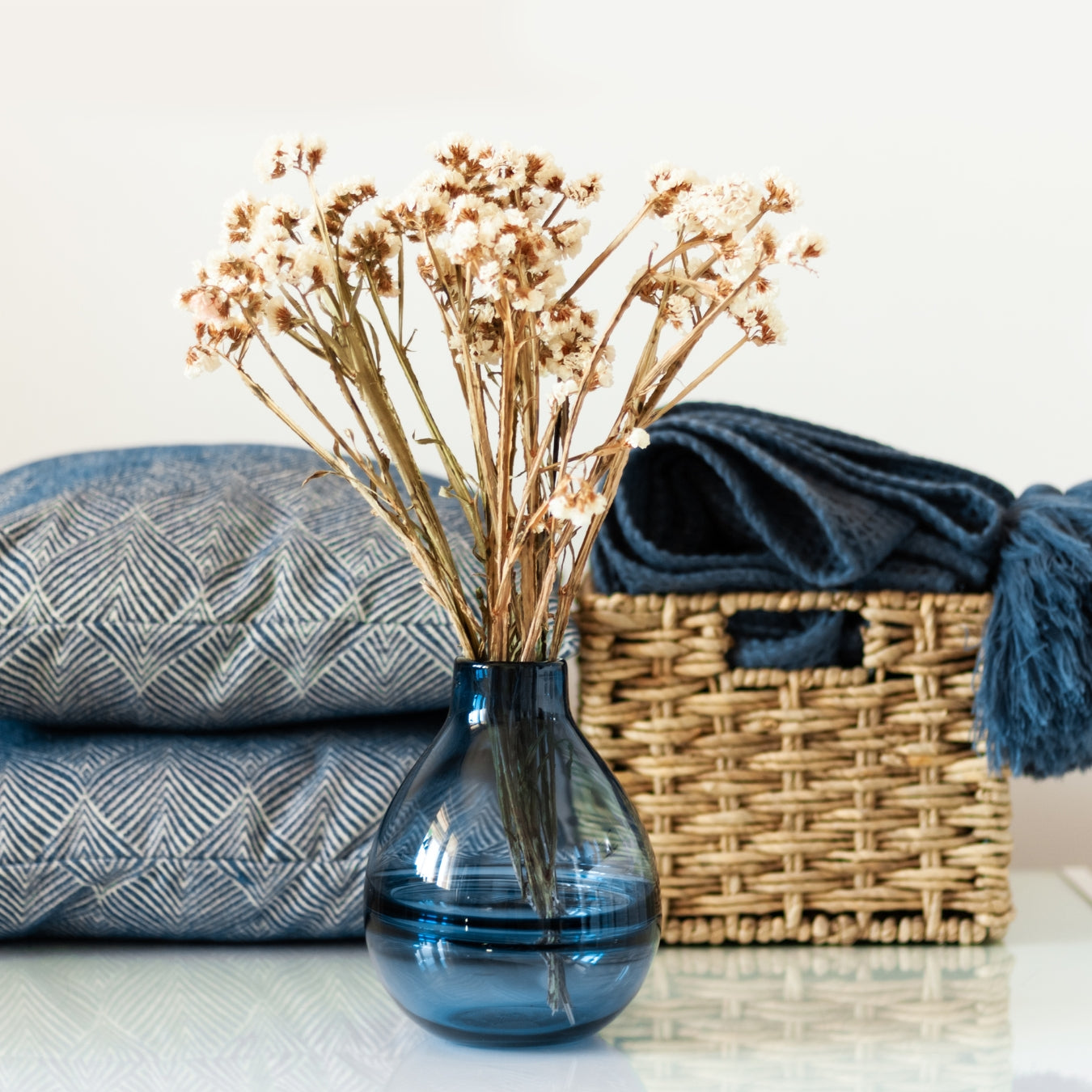 Blue vase with dried flowers on a surface with pillows and a basket in the background