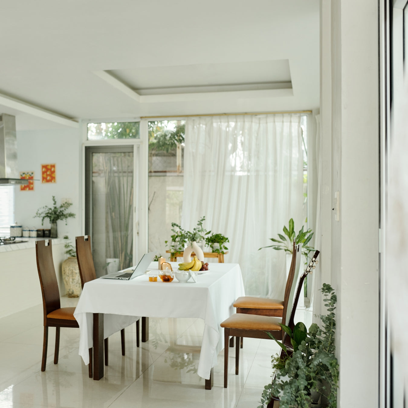 Dining area with a table set for a meal, surrounded by chairs and plants in a bright room.